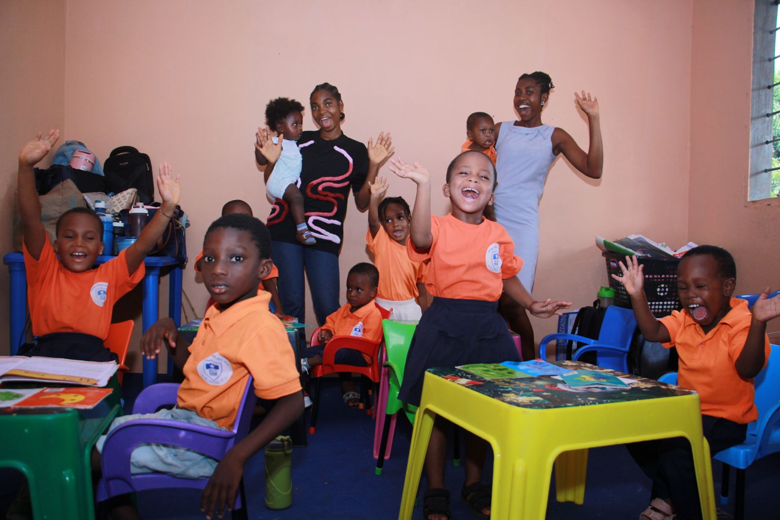Children in orange Divine Favour uniforms waving and laughing in their classroom with two of their teachers